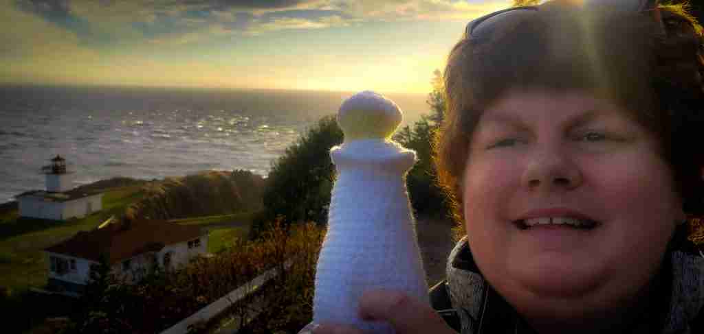 Nancy holding a knitted lighthouse in a selfie with the Cape D'Ors lighthouse in the background.